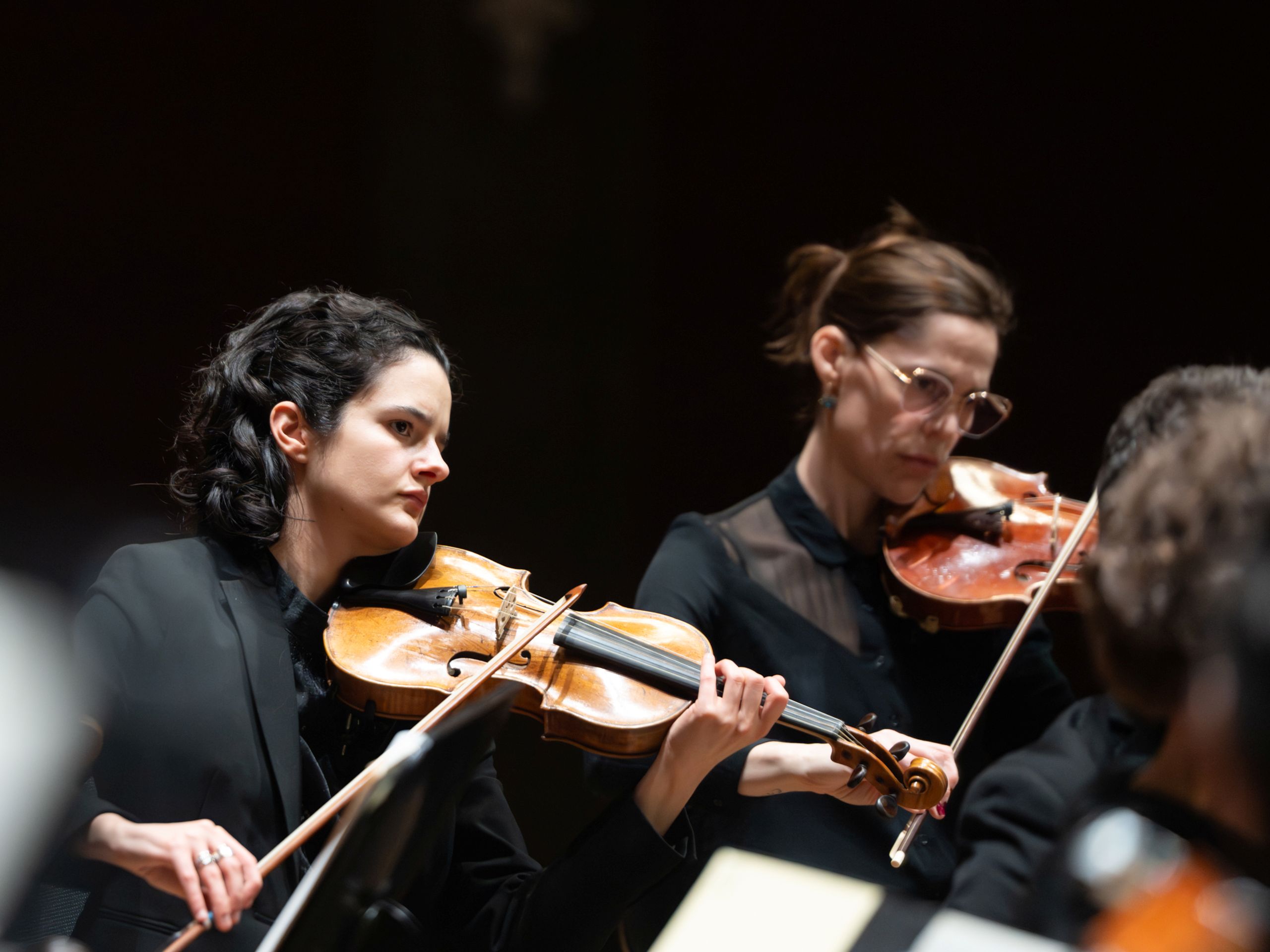 Zwei Orchestermusikerinnen spielen auf ihren Geigen. Sitzend im Orchester.