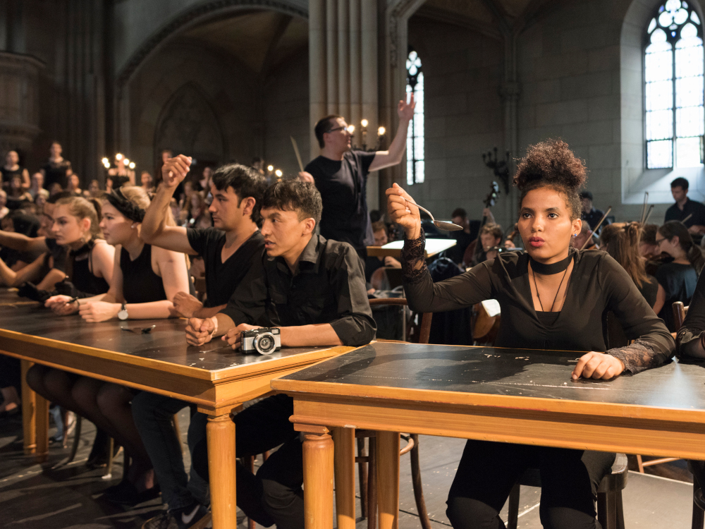 Saenger:innen singen an Tischen, Orchester im Hintergrund