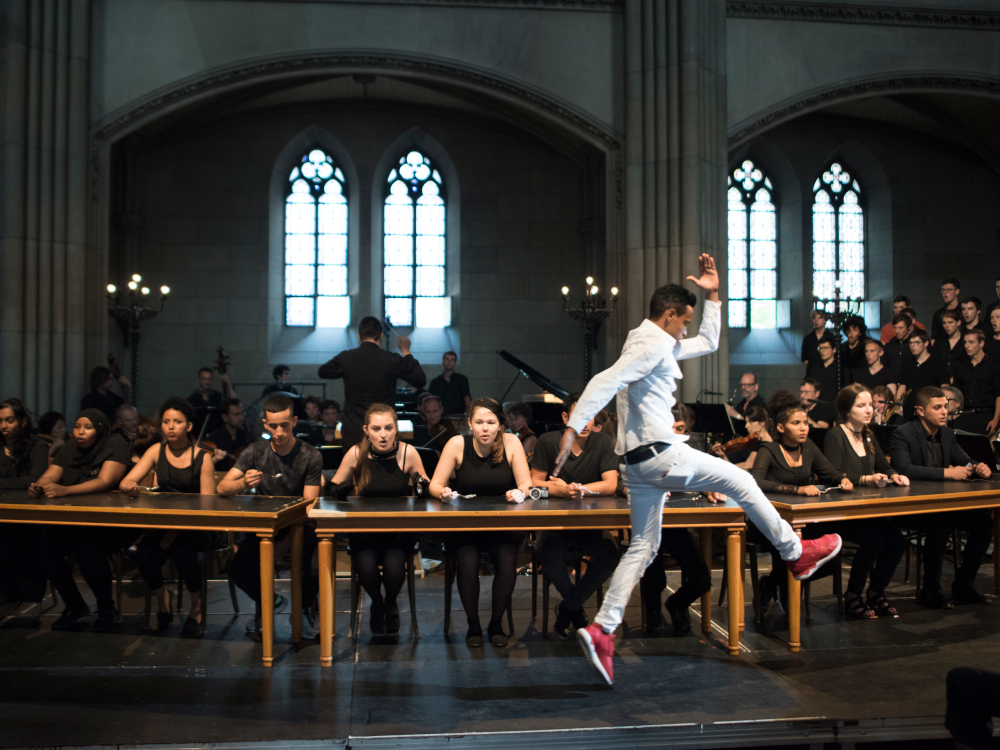 Orchester und Saenger:innen im Hintergrund, einzelner Darsteller springend im Vordergrund