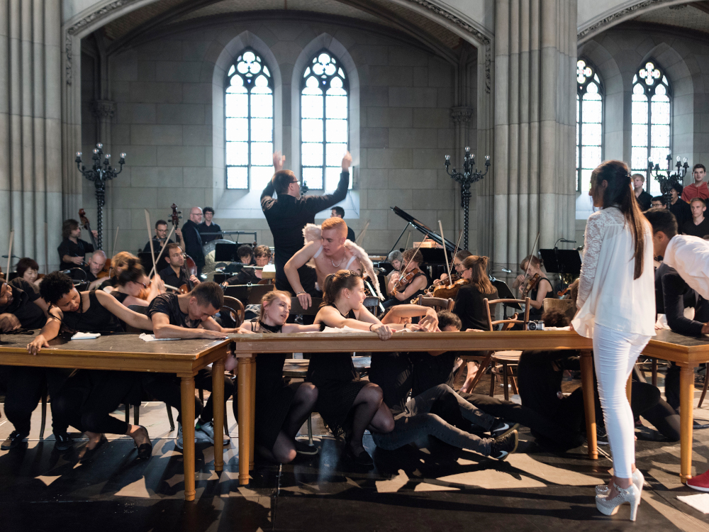 Saenger:innen um Tische gruppiert, Orchester im Hintergrund