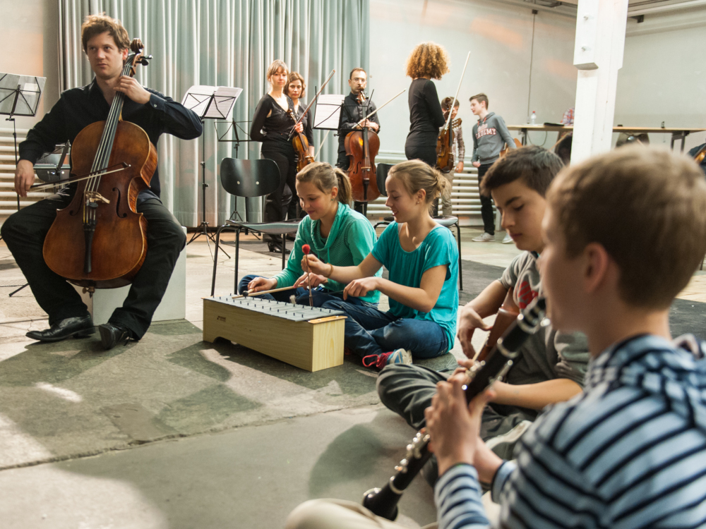 Kinder und ein Cellist musizieren im Vordergrund, Orchestermusiker:innen stehen im Hintergrund