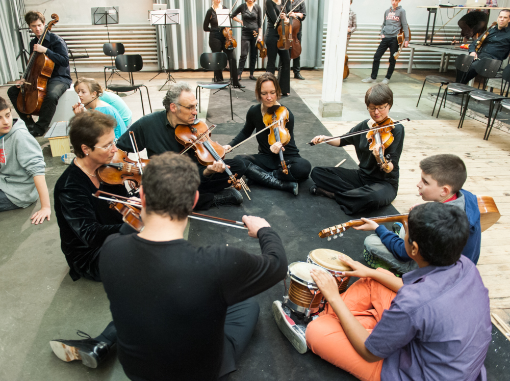 Kinder und Orchestermusiker:innen sitzen musizierend auf dem Boden in einem Kreis
