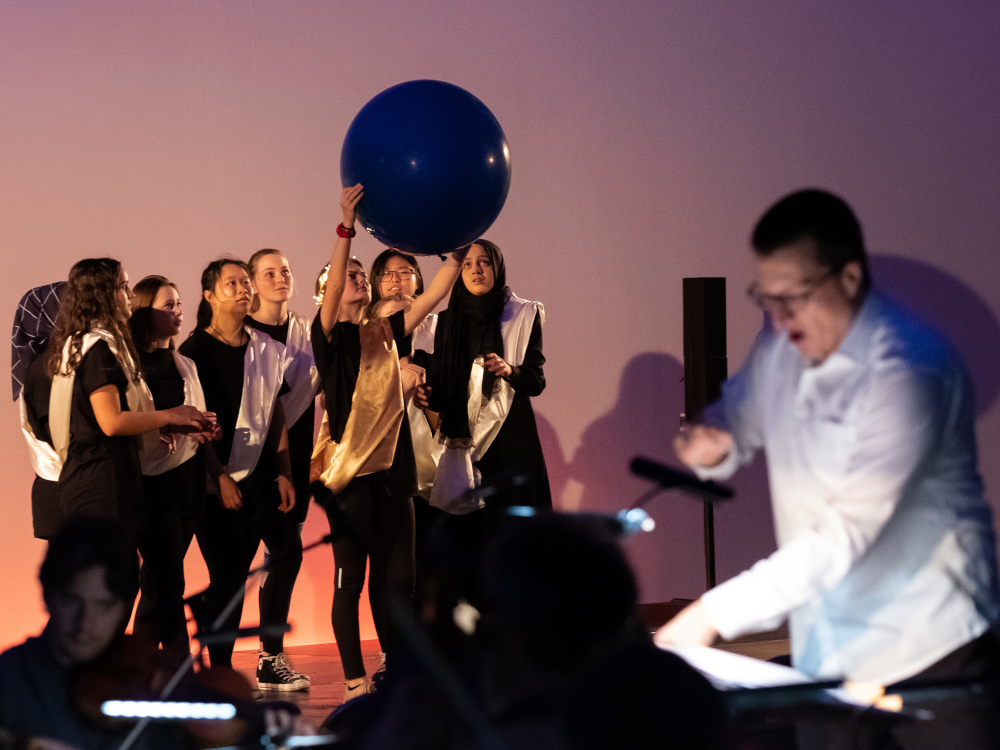 junge Darstellerinnen zu einem grossen Ball hingewandt, im Vordergrund schemenhaft der Dirigent