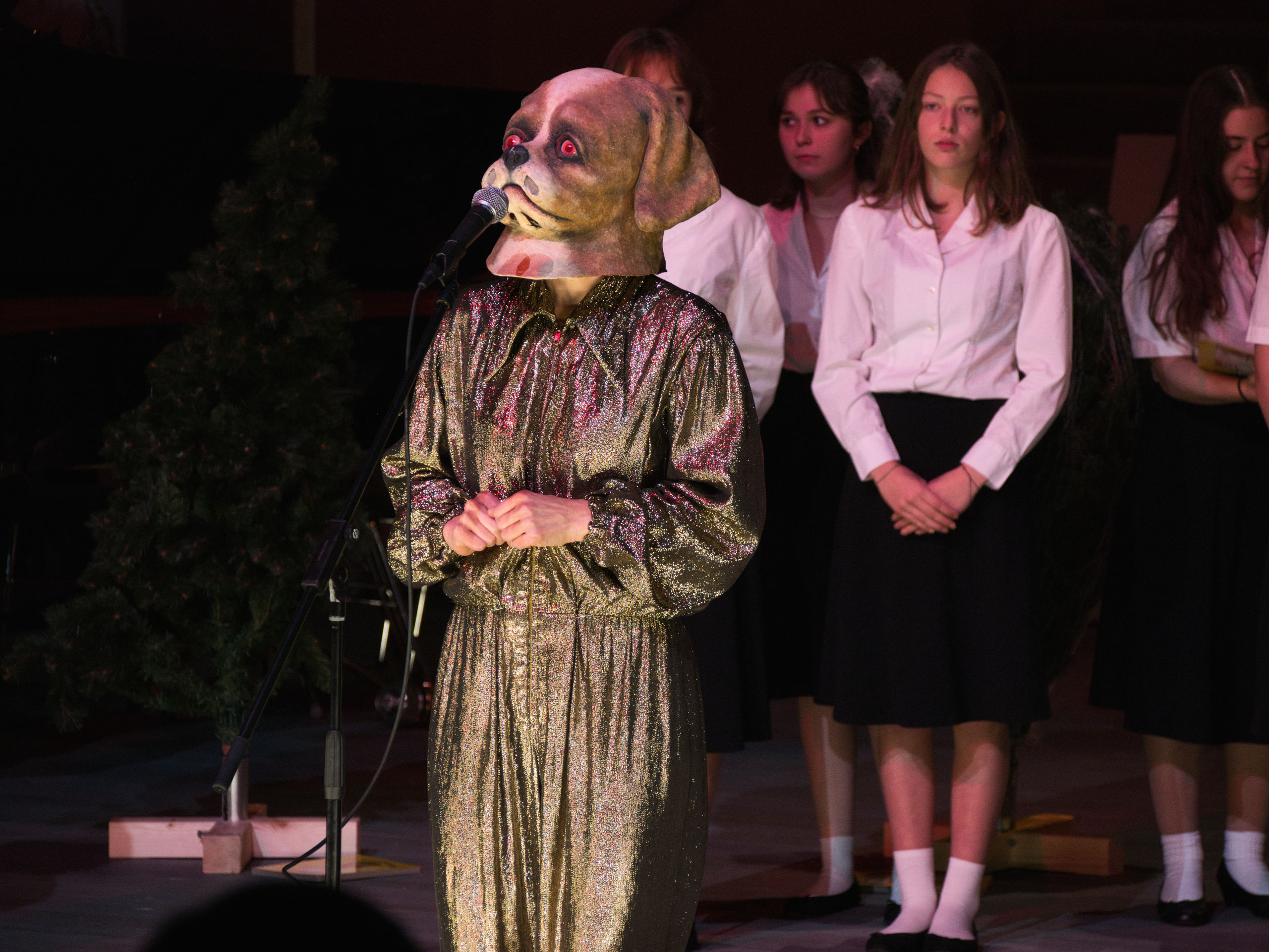Eine als Hund verkleidete Figur spricht in ein Mikrofon. Im Hintergrund: Junge Frauen in Konzertkleidung