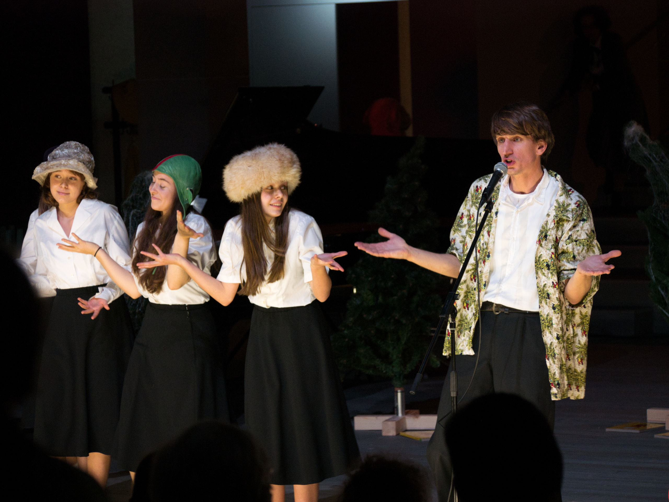 Vier Personen stehen in Konzertkleidung und mit Hüten auf dem Kopf in einer Reihe. Sie gestikulieren und singen.