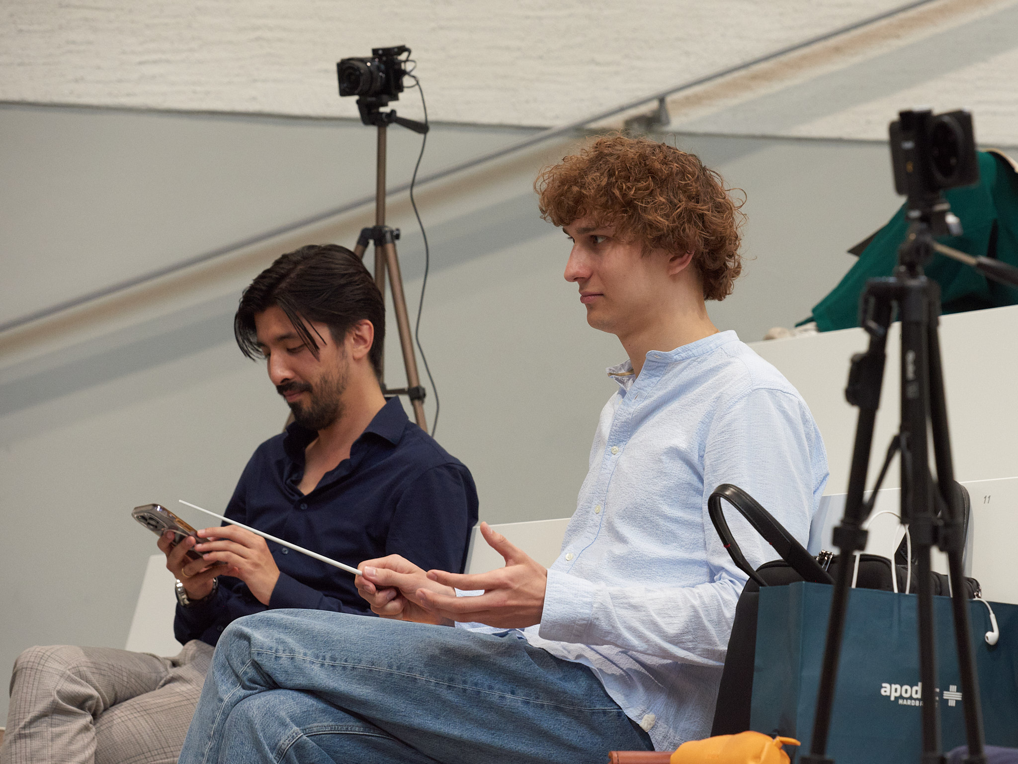 Zwei Junge Herren sitzen auf einer Bank, einer mit einem Dirigierstab, einer mit dem Handy in der Hand.