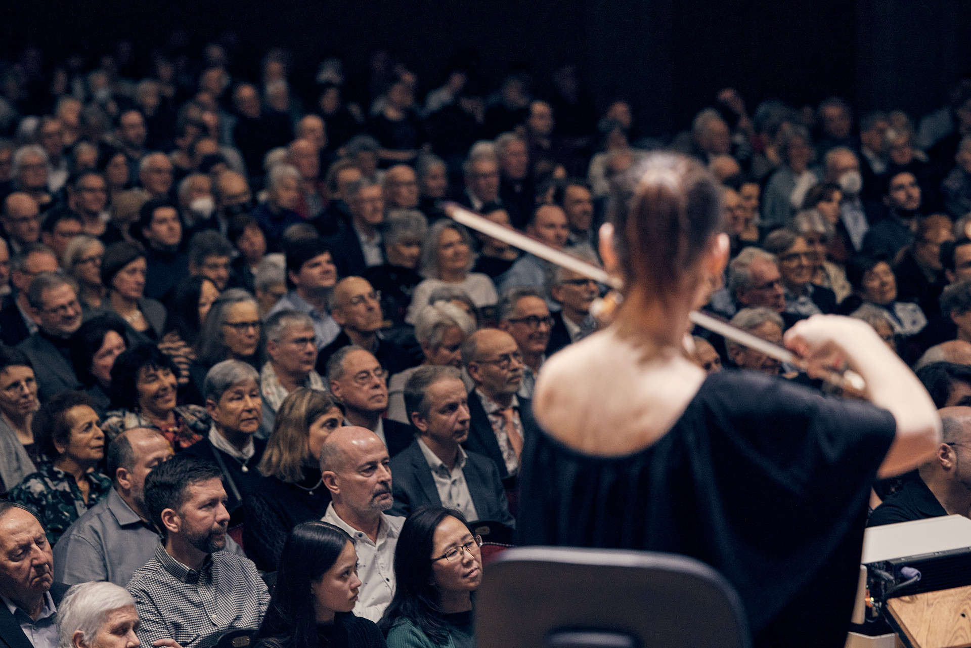 Blick von der Buehne ins Publikum. Im Vordergrund der Ruecken einer Geigerin.