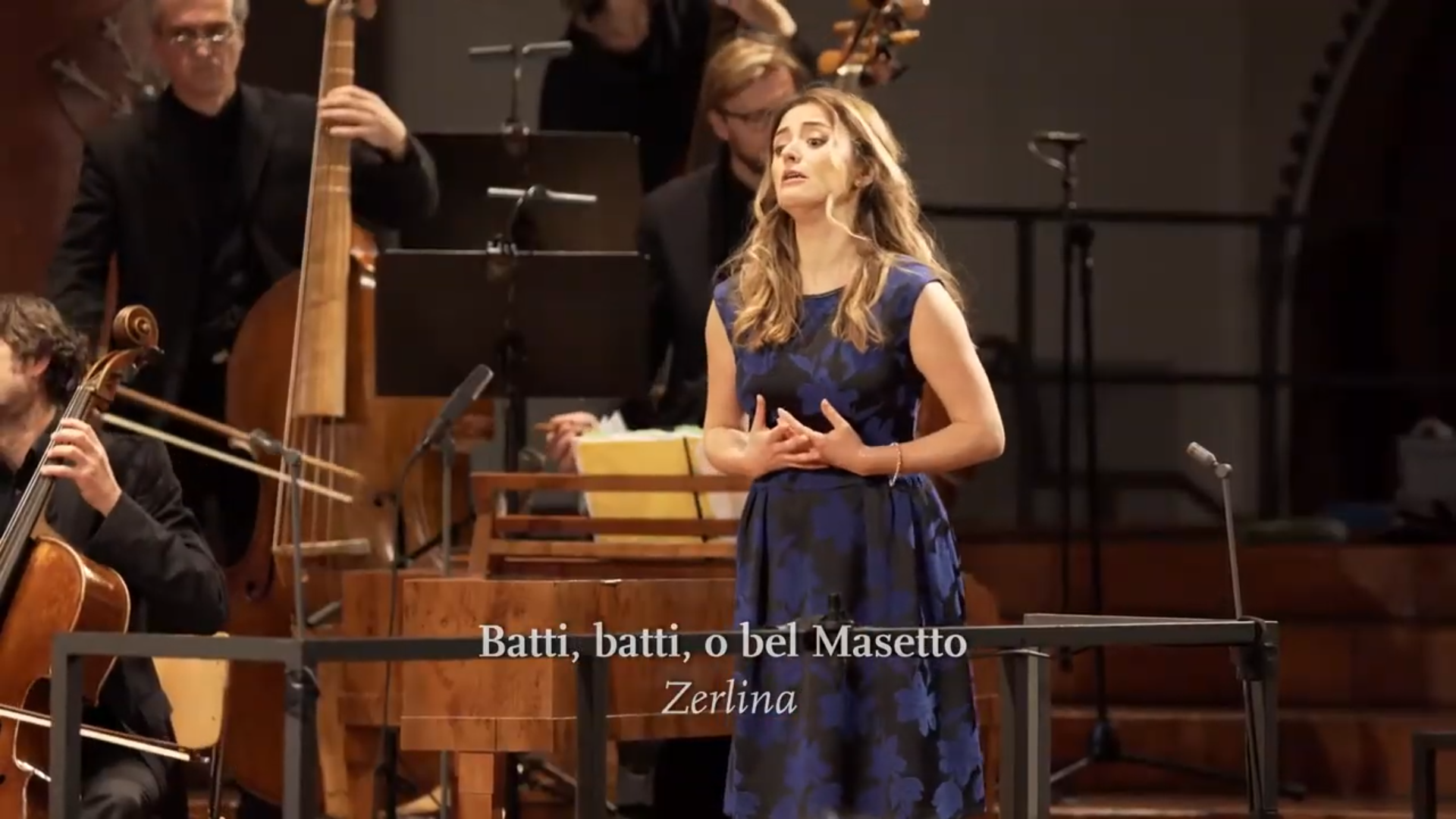 Eine Solistin singt in einer Kirche, im Hintergrund sind einige Orchestermusiker zu sehen. Eingeblendet ist der Titel "Batti, batti, o bel Masetto".