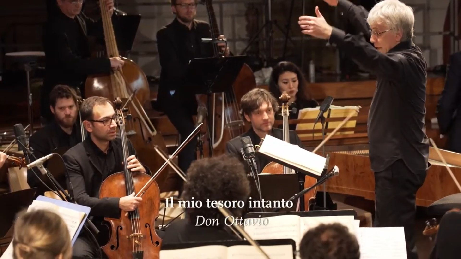Der Dirigent Giovanni Antonini beim Dirigieren, ein Teil der Streichersektion des Orchesters ist zu sehen. Eingeblendet ist der Titel "Il mio tesoro intanto".