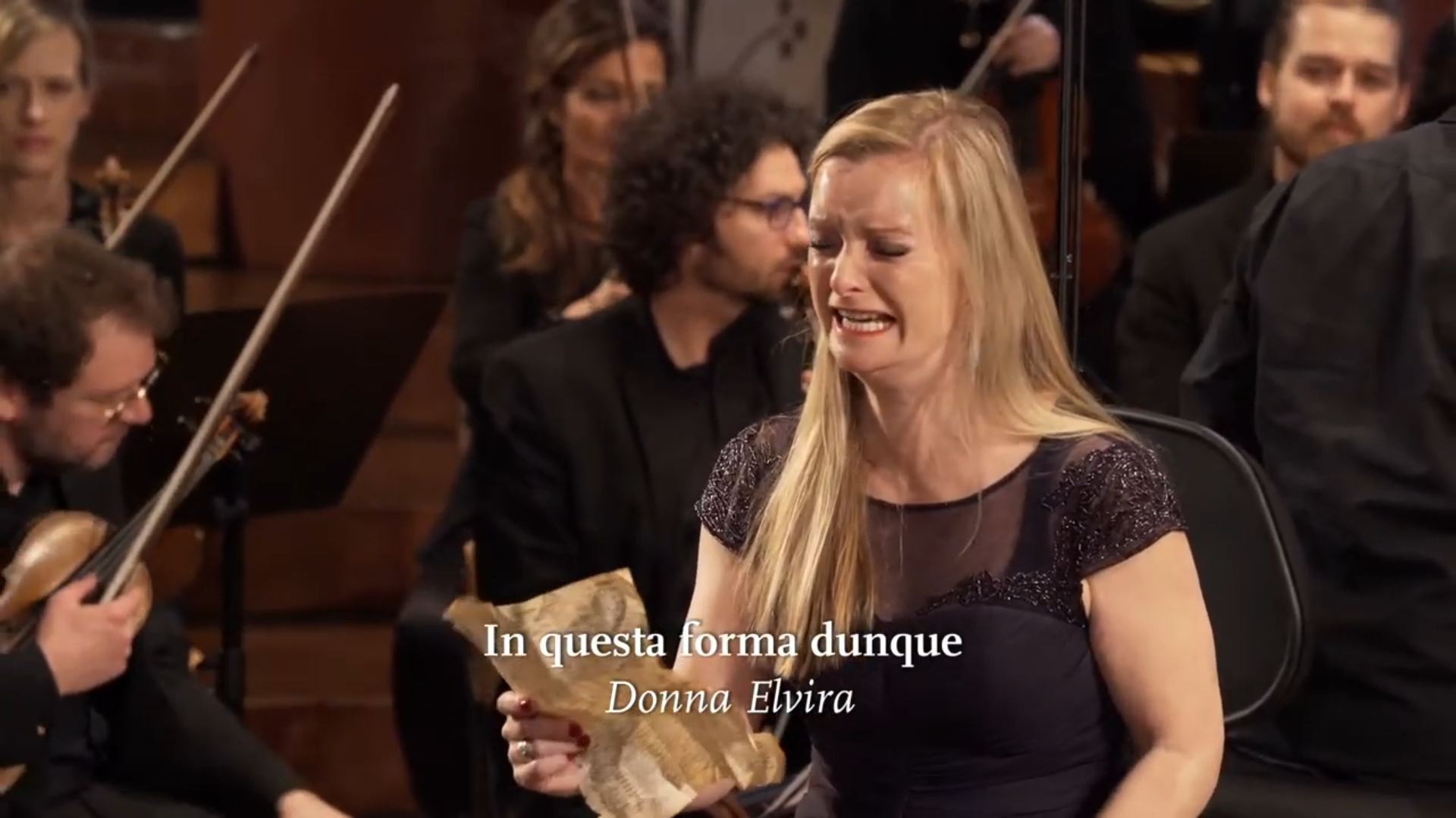 Eine Solistin singt mit schmerzhaft verzogenem Gesicht, im Hintergrund wartet das Orchester auf seinen Einsatz. Eingeblendet ist der Titel "In questa forma dunque".