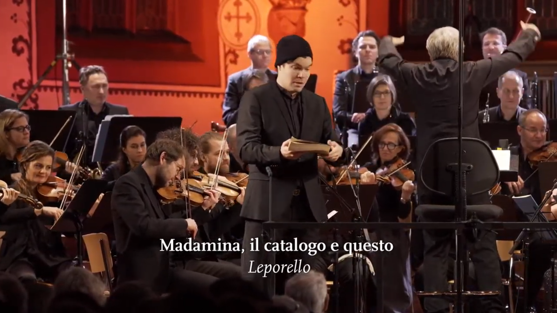 Ein Solist, mit einer Mütze und einem dicken Buch ausgestattet, singt im Vordergrund, im Hintergrund spielt das Orchester . Eingeblendet ist der Titel "Madamina, il catalogo e questo".
