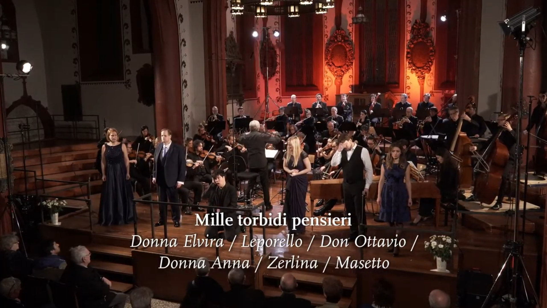 Das Kammerorchester Basel spielt in einer Kirche, im Vordergrund singen sechs Solist:innen, einer davon am Boden kniend. Eingeblendet ist der Titel "Mille torbidi pensieri".