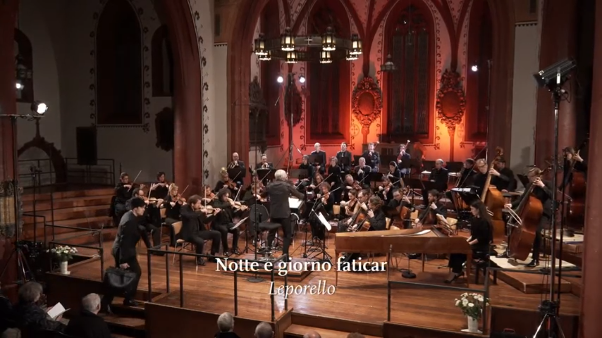 Das Kammerorchester Basel spielt in einer Kirche, im Vordergrund betritt ein Solist mit Hut und Aktenkoffer die Bühne. Eingeblendet ist der Titel "Notte e giorno faticar".