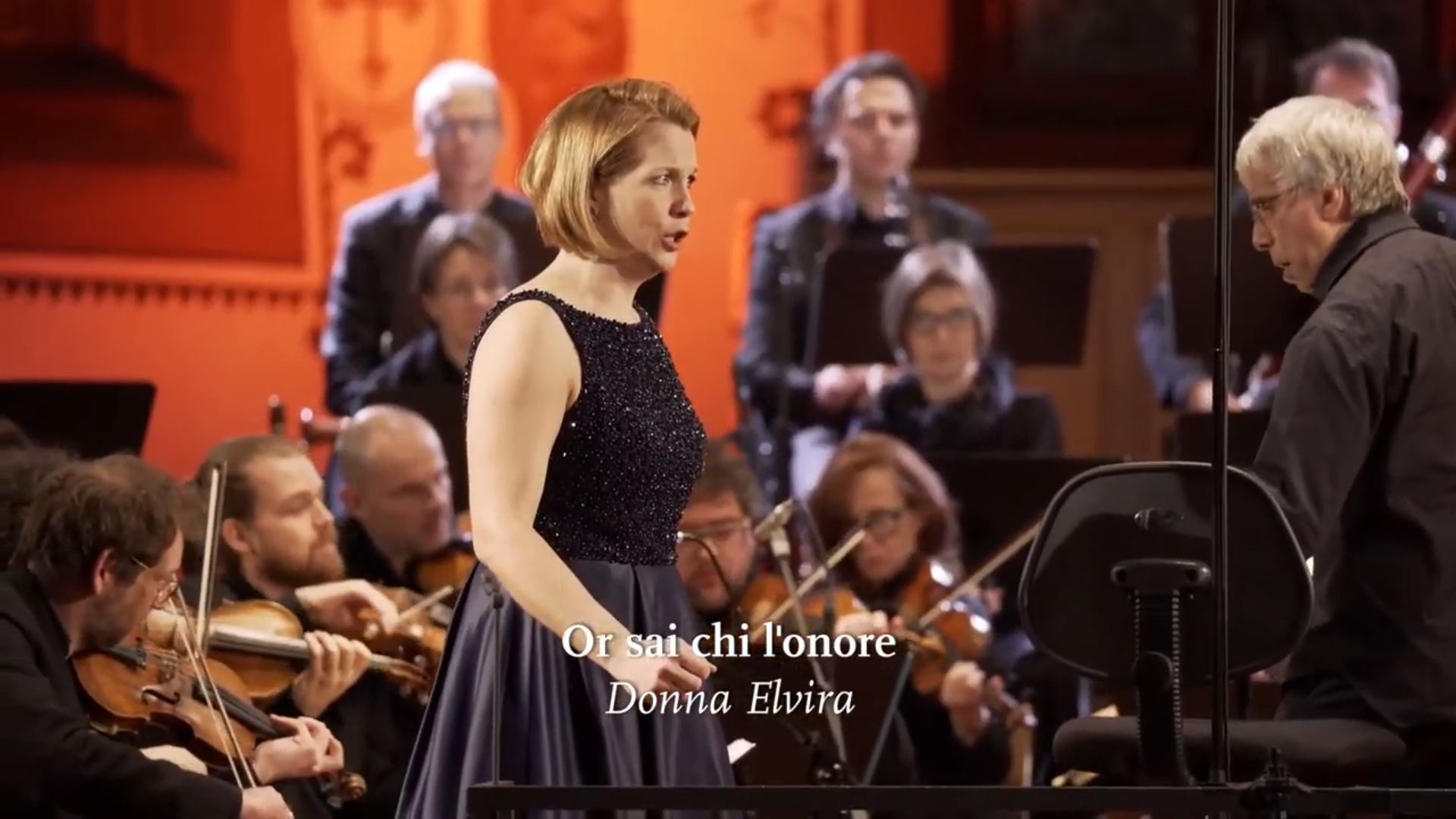 Eine Solistin singt seitlich zum Dirigenten Giovanni Antonini gewendet, im Hintergrund spielt das Orchester. Eingeblendet ist der Titel "Or sai chi l'onore".