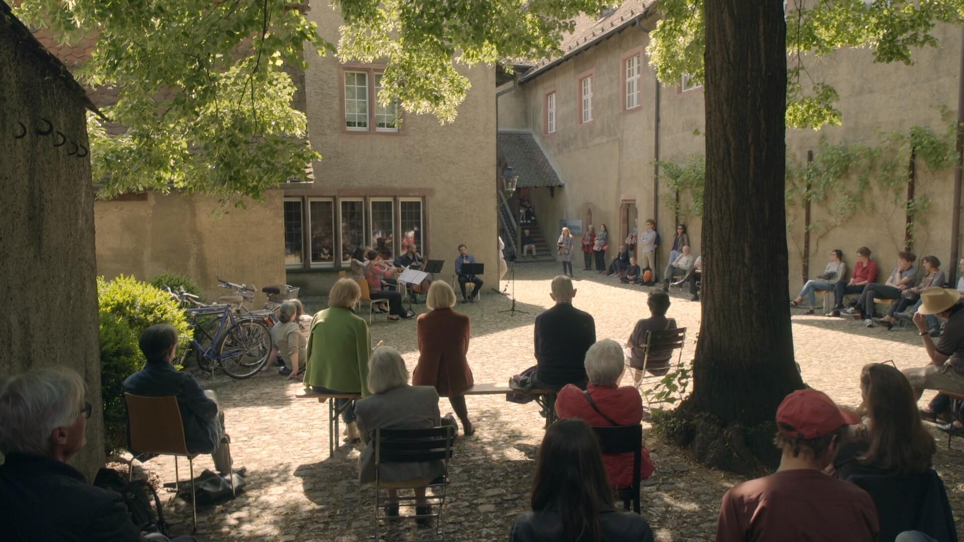 Musiker:innen geben ein Konzert in einem Innenhof. Das Publikum sitzt oder steht im Hof verteilt.