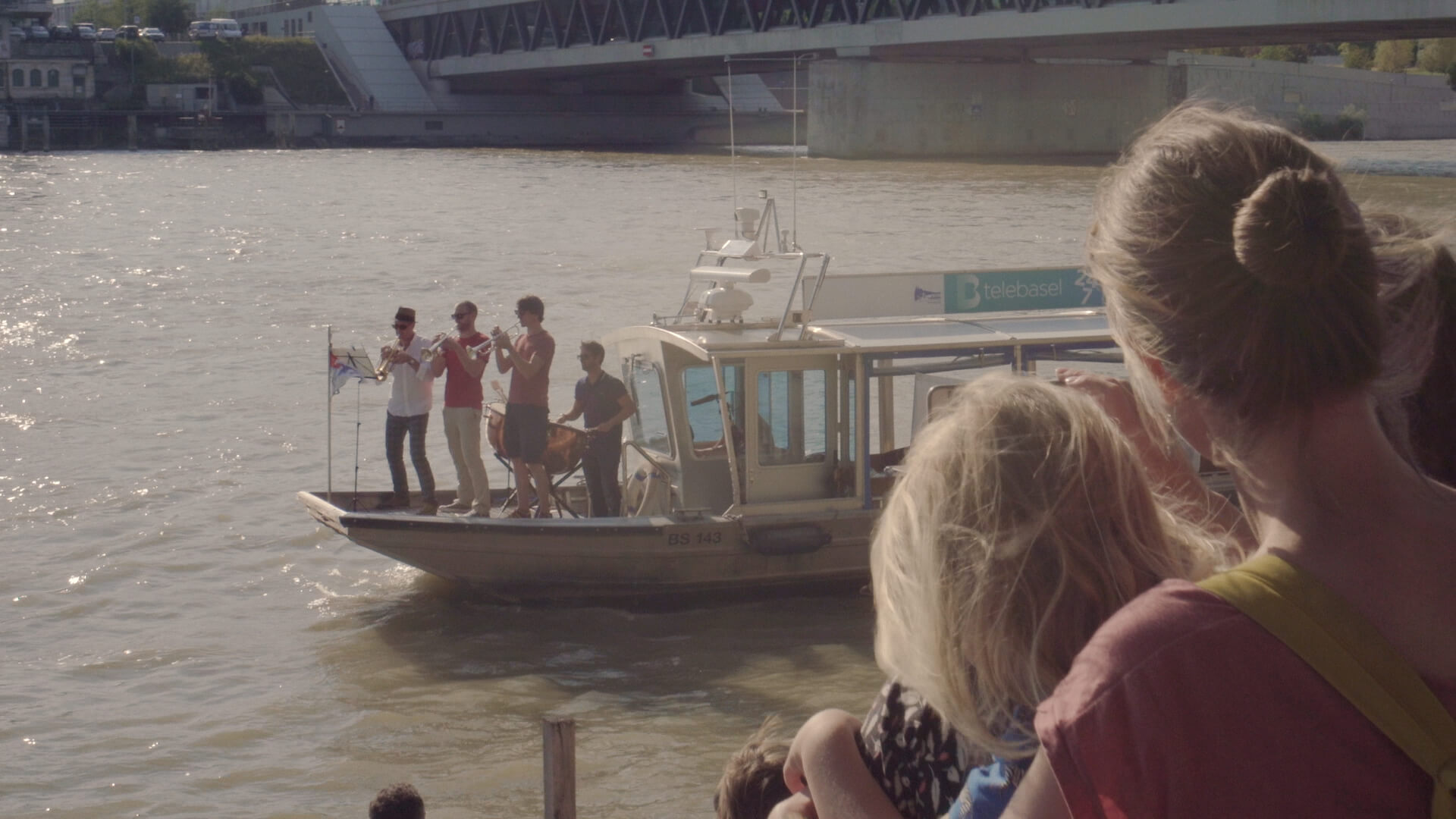 Drei Trompeter und ein Perkussionist spielen vorne auf einem Boot, das auf dem Rhein schwimmt. Publikum hört vom Ufer aus zu.