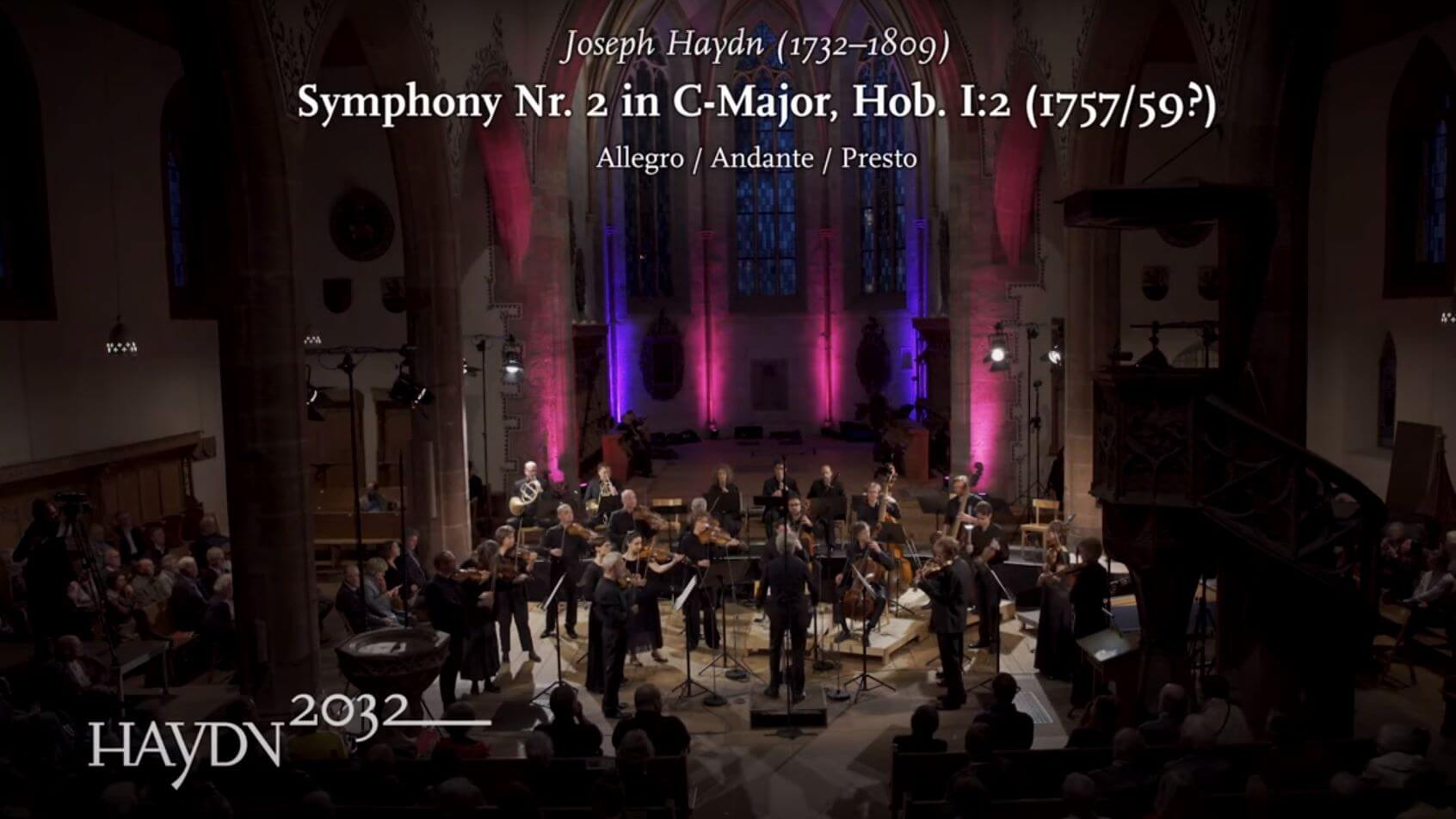 Das Kammerorchester Basel spielt in einer Kirche. Eingeblendet ist der Text: Joseph Haydn, Symphony No. 2 in C-Major, Hob. I:2 und das Logo des Projekts Haydn2032.