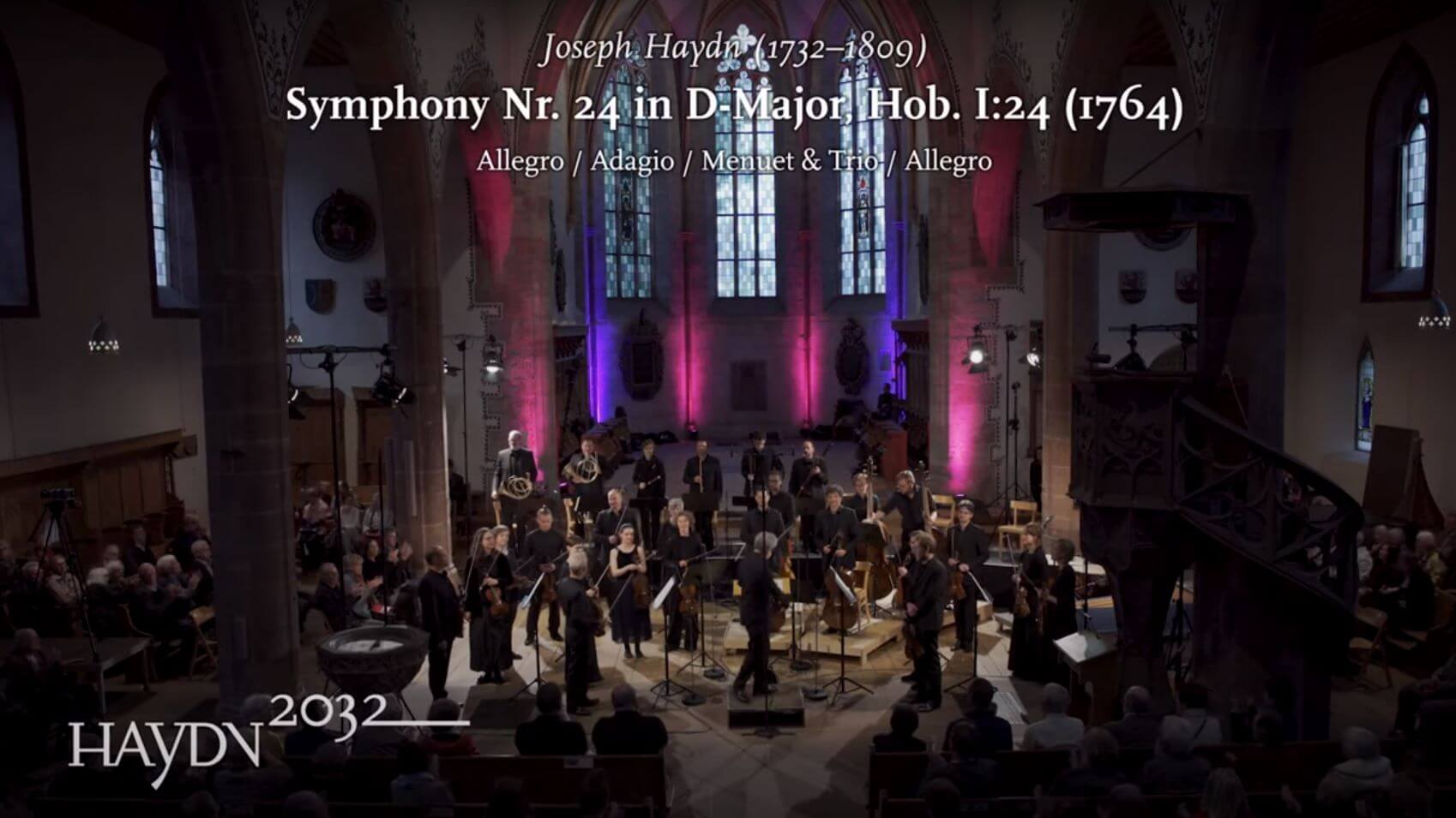Das Kammerorchester Basel spielt in einer Kirche. Eingeblendet ist der Text: Joseph Haydn, Symphony No. 24 in D-Major, Hob. I:24 und das Logo des Projekts Haydn2032.