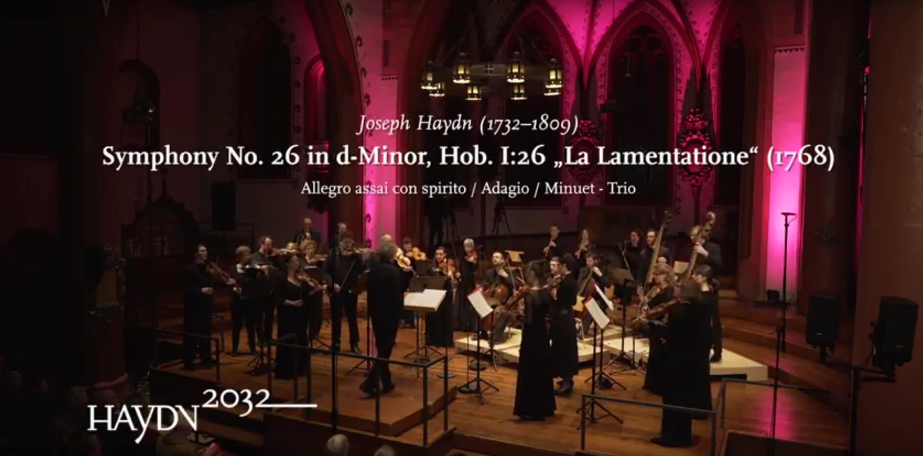 Das Kammerorchester Basel spielt in einer Kirche. Eingeblendet ist der Text: Joseph Haydn, Symphony No. 26 in d-Minor, Hob. I:26 "La Lamentatione" (1768) und das Logo des Projekts Haydn2032.