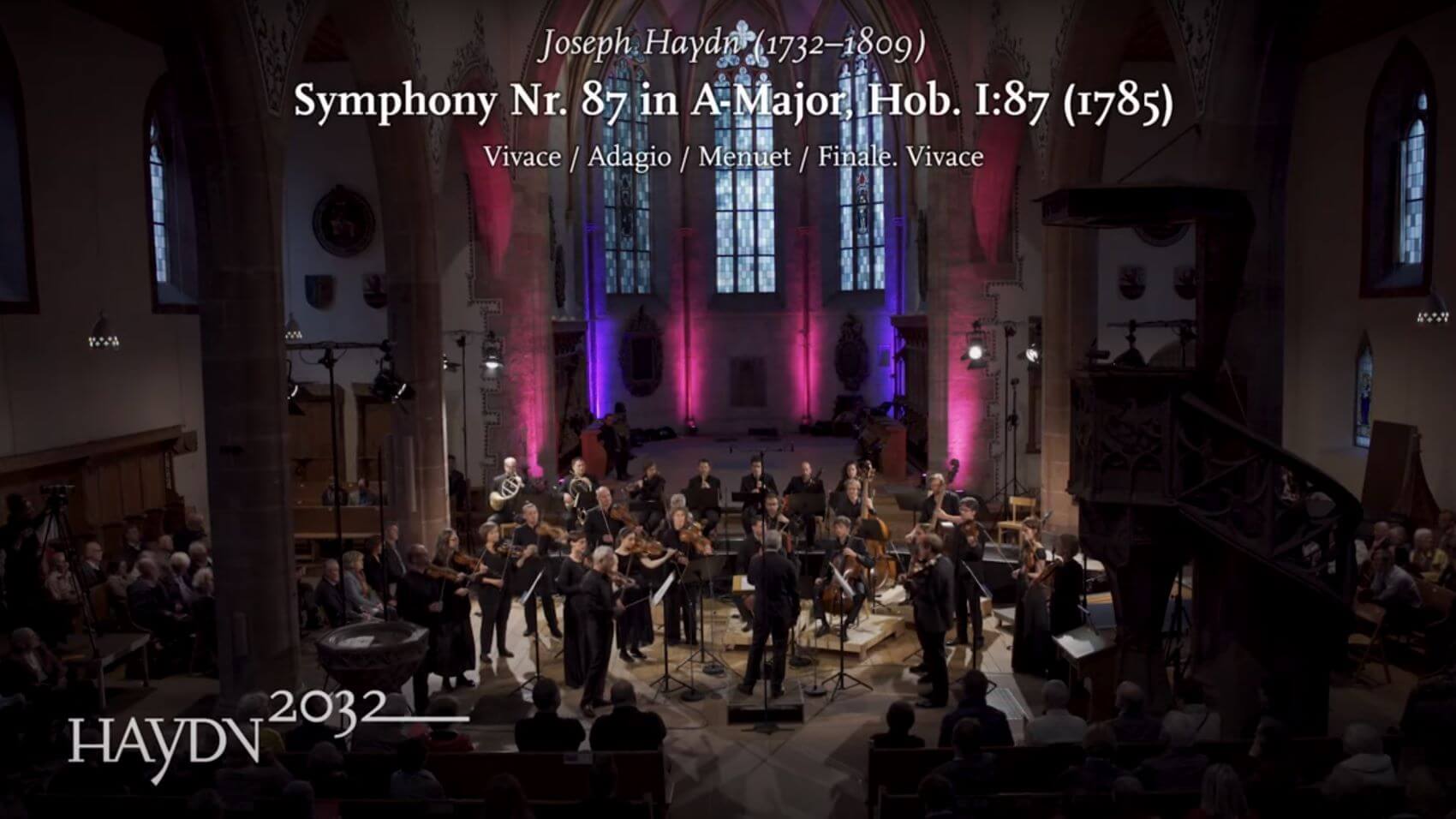 Das Kammerorchester Basel spielt in einer Kirche. Eingeblendet ist der Text: Joseph Haydn, Symphony No. 87 A-Major, Hob. I:87 und das Logo des Projekts Haydn2032.