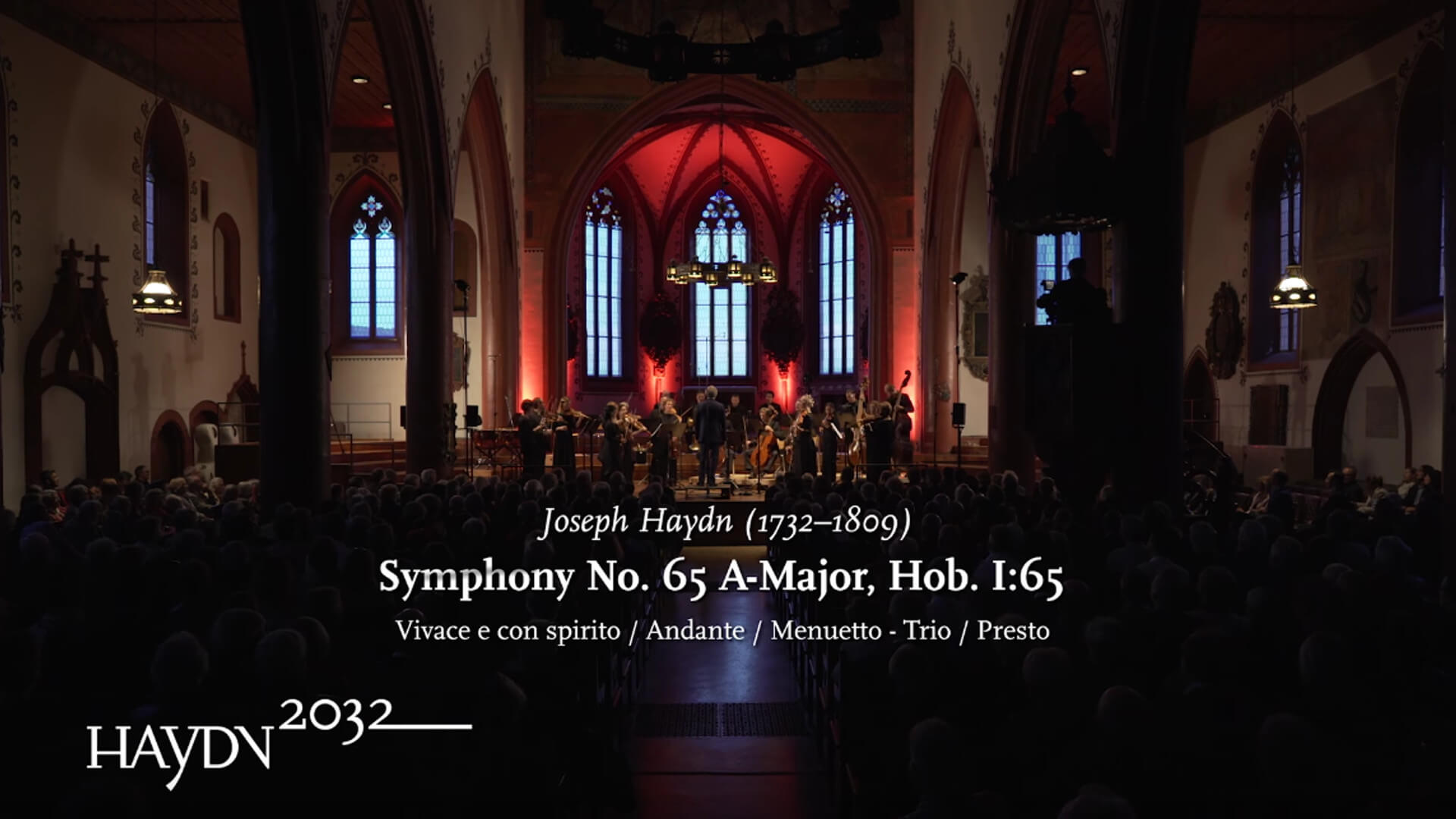 Das Kammerorchester Basel spielt in einer Kirche. Eingeblendet ist der Text: Joseph Haydn, Symphony No. 65 A-Major, Hob. I:65 und das Logo des Projekts Haydn2032.