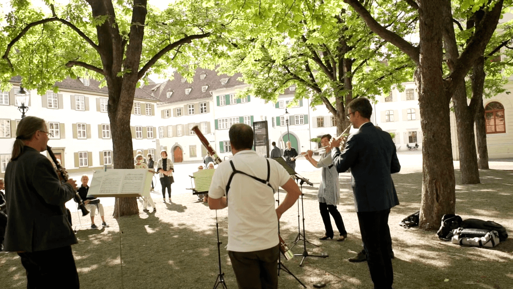 Musiker:innen des Orchesters (Flöte, Horn, Fagott, Klarinette) spielen unter Bäumen auf dem Münsterplatz in Basel.