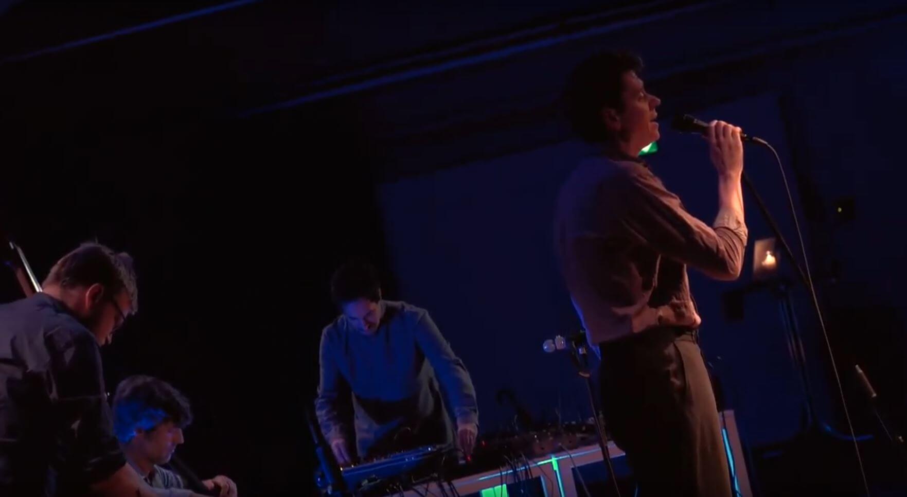 Konzertraum in blau beleuchtet, ein Sänger steht im Zentrum, daneben eine Person am Mischpult.