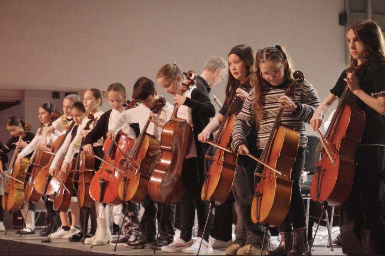 Kinder stehen in einer Reihe auf der Bühne und spielen Cello.
