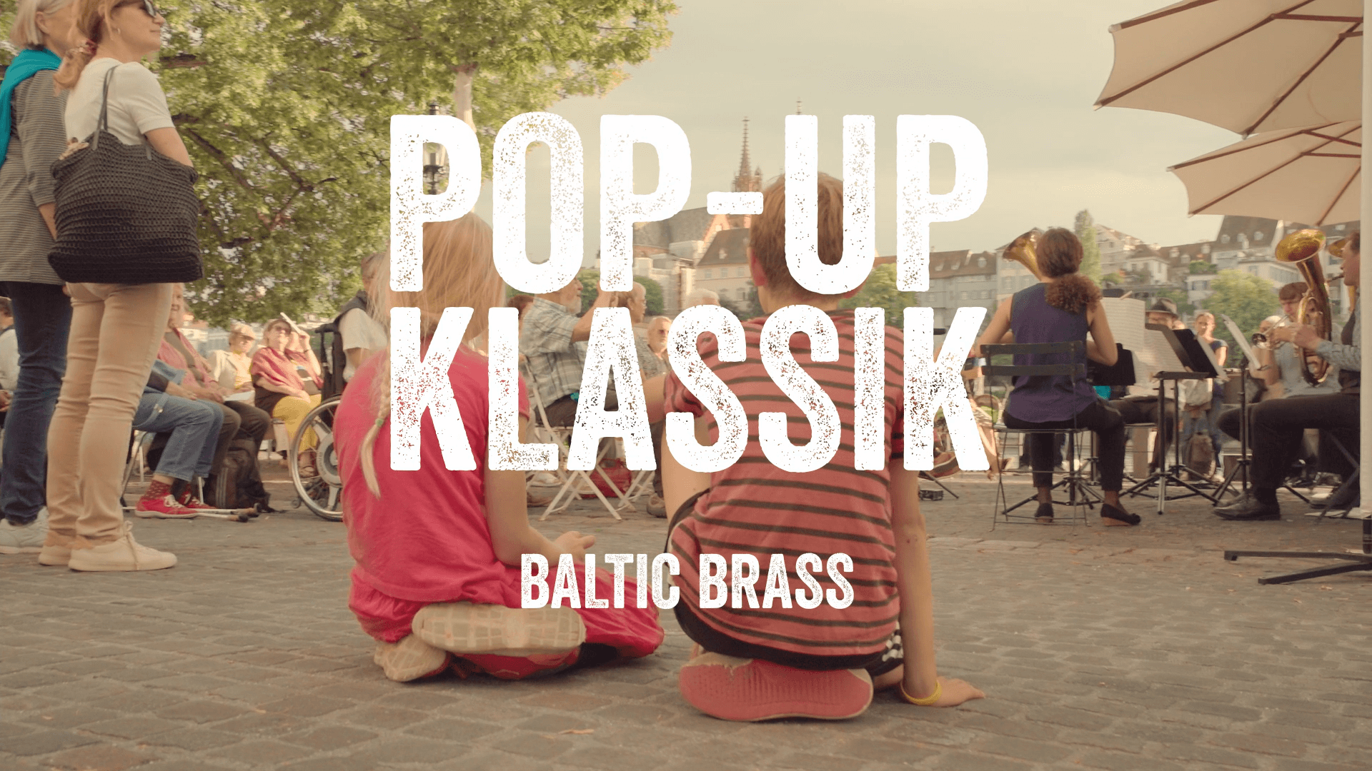 Zwei Mädchen sitzen in der Stadt auf dem Boden und lauschen einem Konzert. Eingeblendet der Text: Pop-Up Klassik, Baltic Brass.