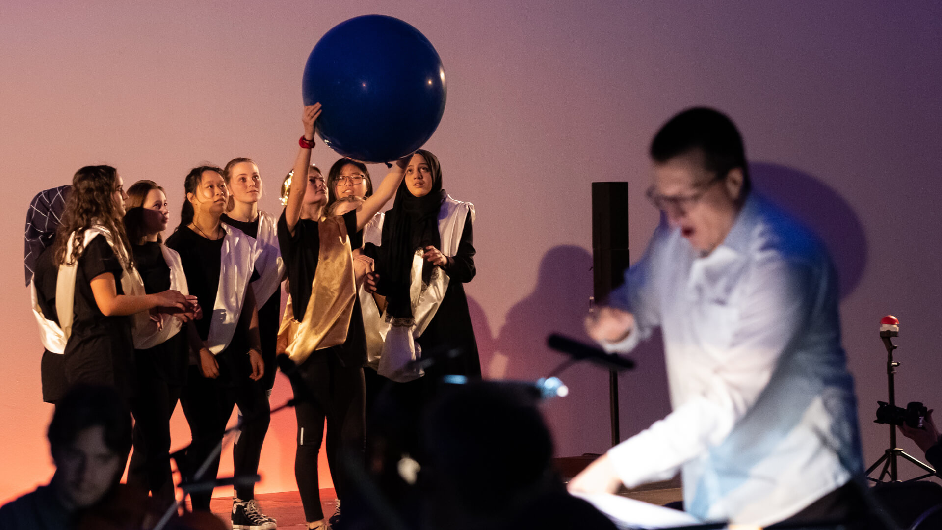Eine Gruppe junger Darstellerinnen hält einen grossen Ball in die Luft. Im Vordergrund ist verschwommen der Dirigent zu sehen.