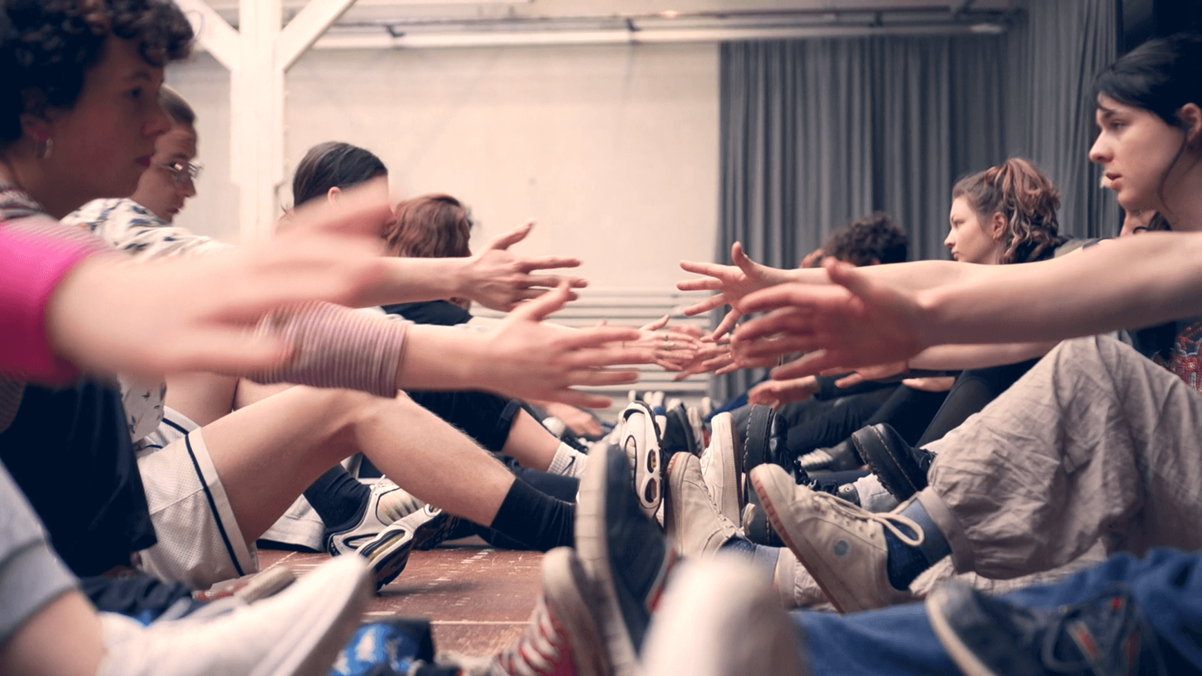 Junge Darsteller:innen sitzen sich am Boden gegenüber und strecken die Arme zueinander hin.