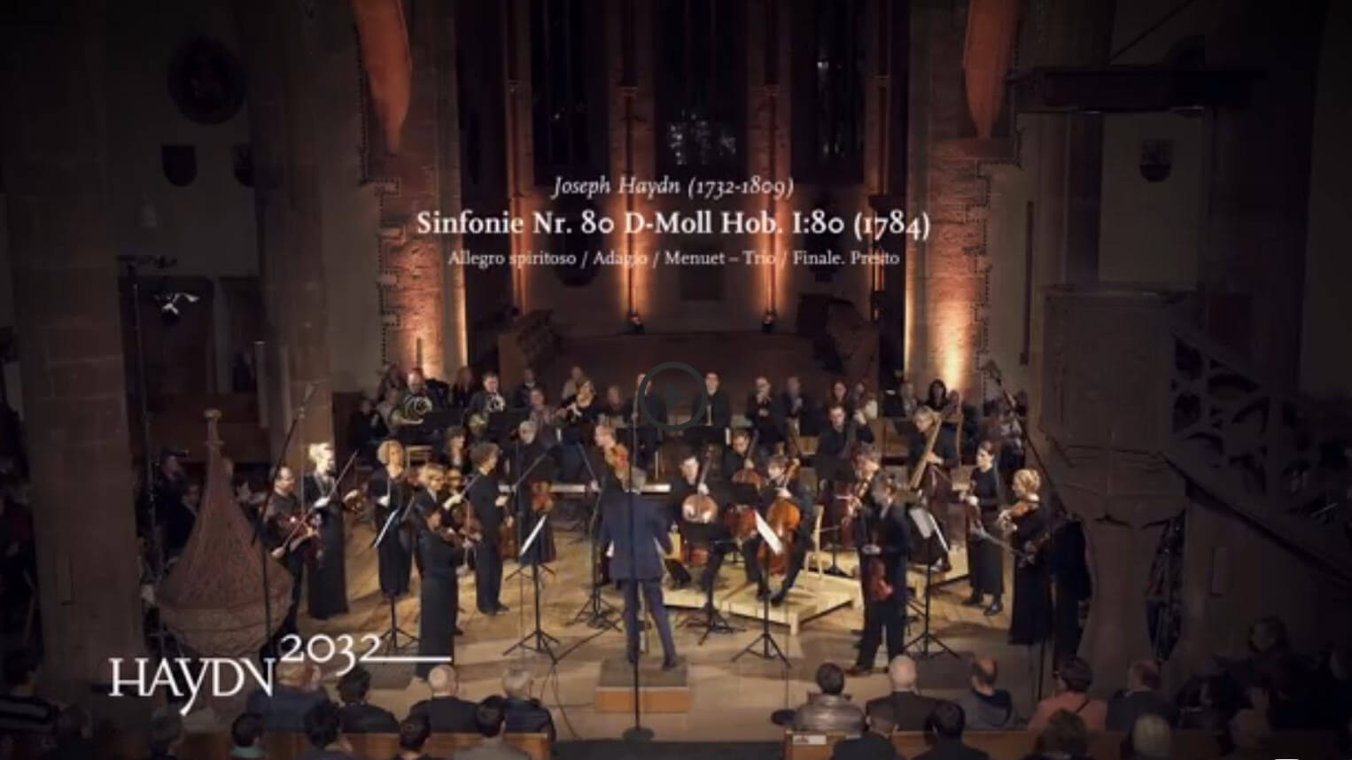Das Kammerorchester Basel spielt in einer Kirche. Eingeblendet ist der Text: Joseph Haydn, Sinfonie Nr. 80 D-Moll Hob. I:80 (1784) und das Logo des Projekts Haydn2032.