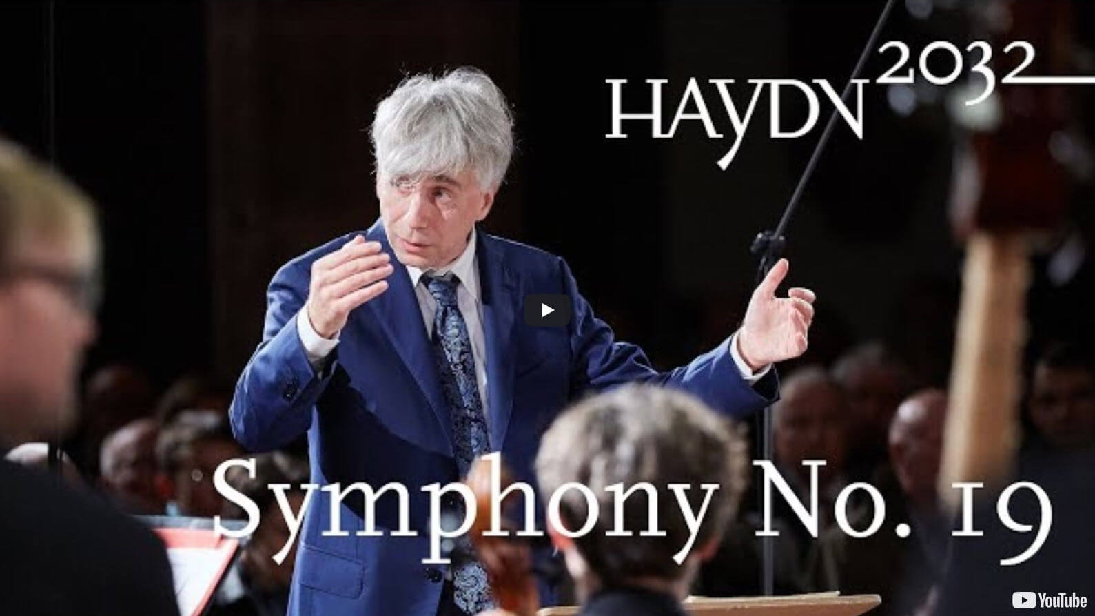 Frontalansicht des Dirigenten Giovanni Antonini beim Dirigieren. Eingeblendet ist der Text "Symphony No. 19" und das Logo des Projekts Haydn2032.